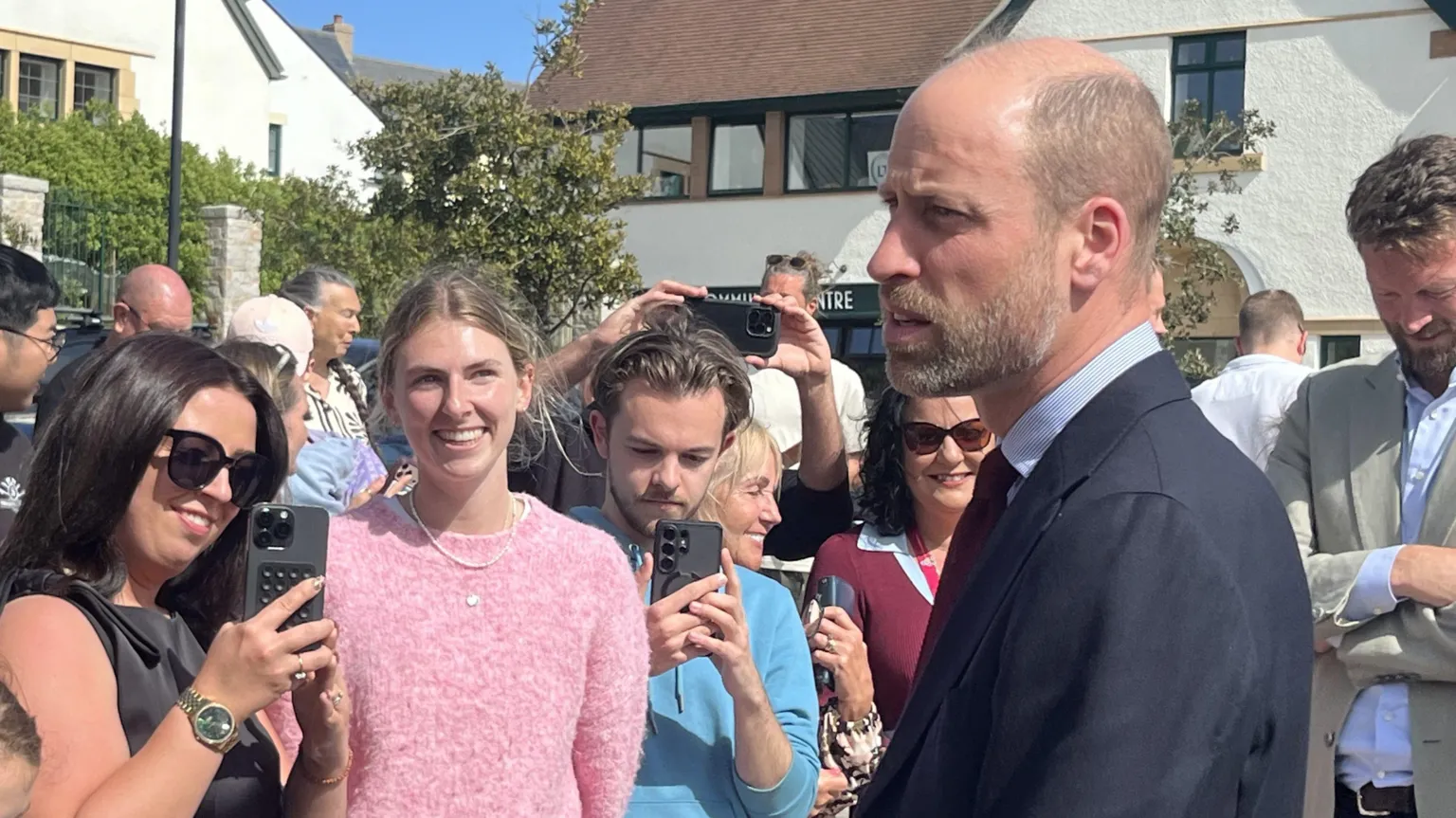 BBC Prince William looking to the left as he talks to a crowd of people. There is a woman smiling in a pink jumper, and a woman with black hair and sun glasses taking a picture. 