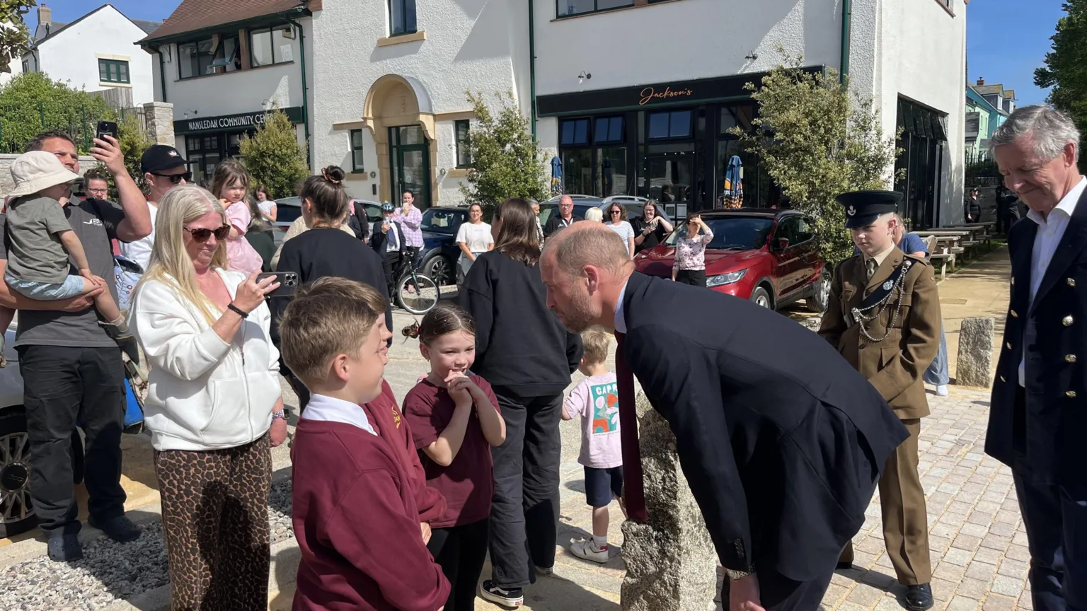 Prince William speaking to school children who are stood in front of a crowd. There's a woman stood behind them taking a photo of them all talking. 